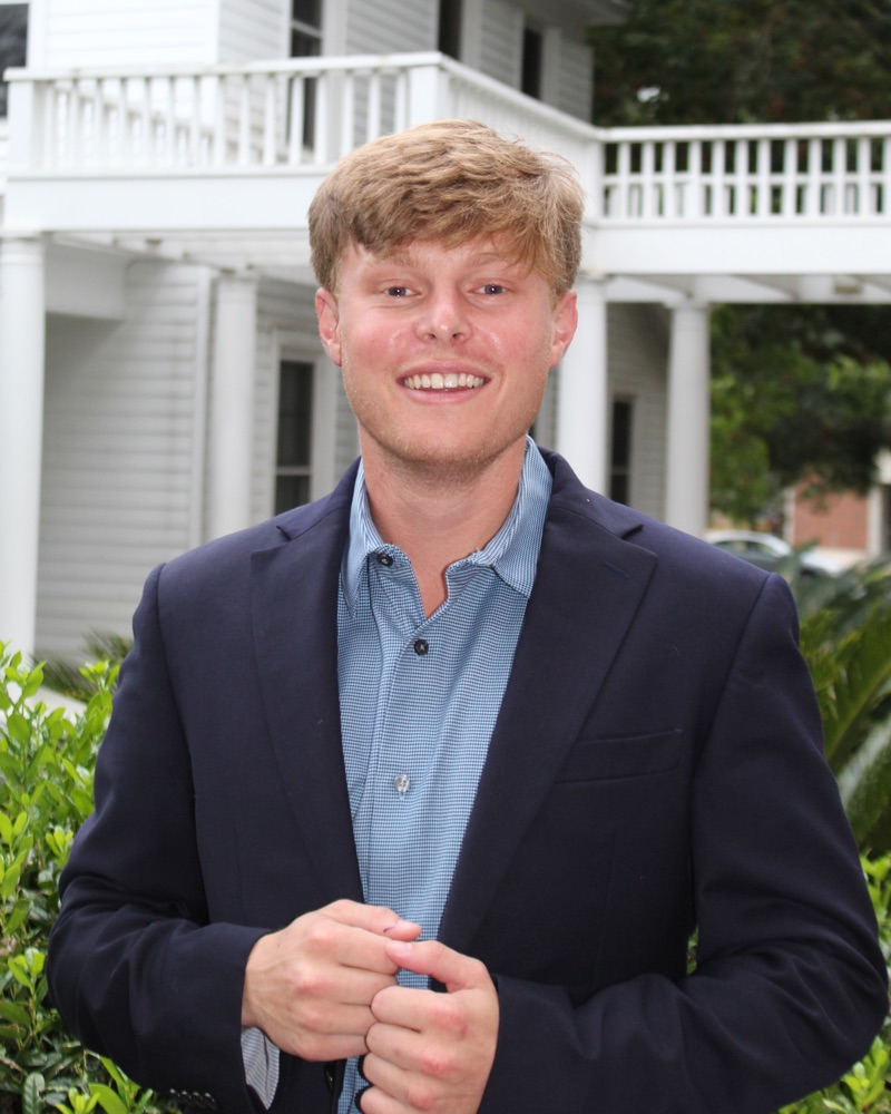 Portrait of Drew M. Solomon smiling, wearing a navy blazer over a light blue collared shirt, photographed outdoors.